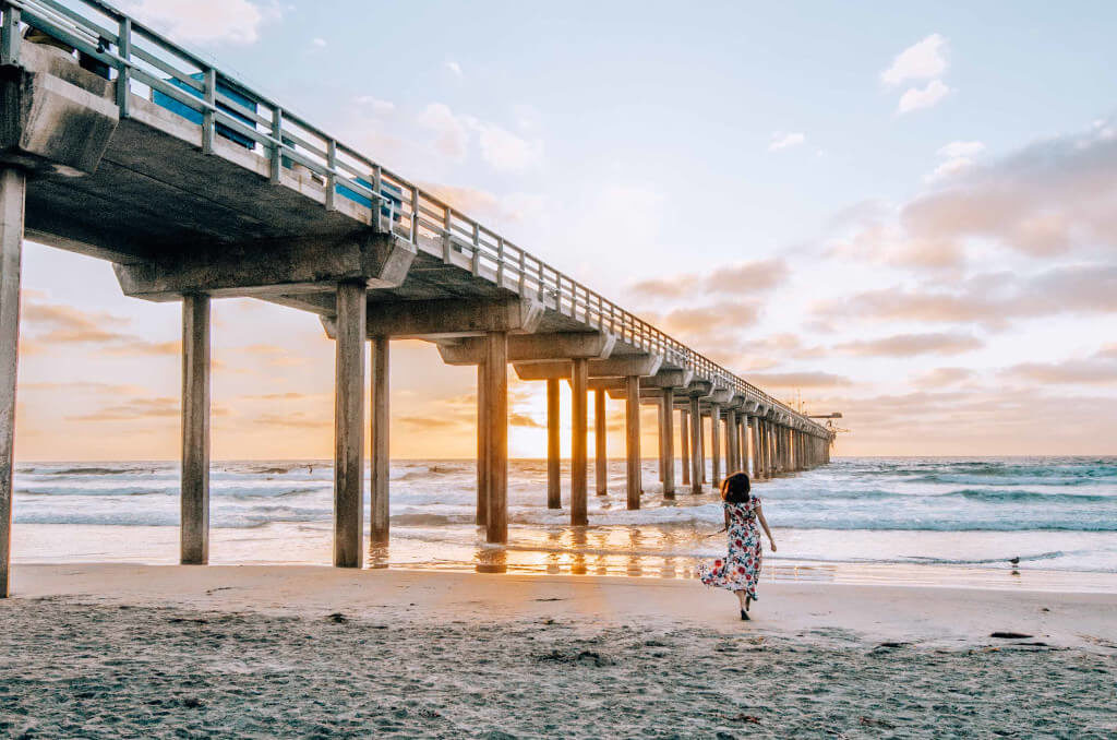Girl running toward the beach with a pier to one side and the pink sunset.