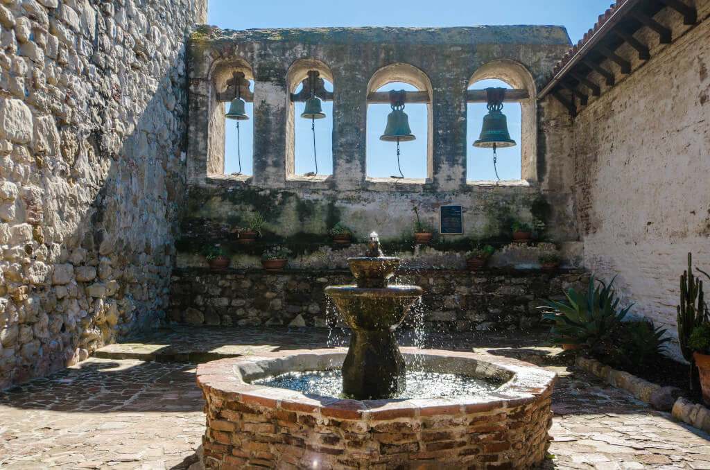 Bells and fountain with the stone walls of Mission San Juan Capistrano