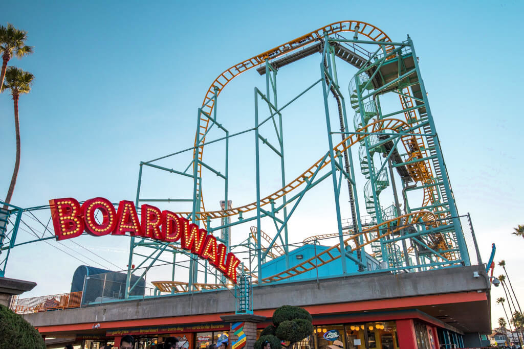Red sign that reads "Boardwalk" with roller coaster and amusement park behind it in Santa Cruz, California