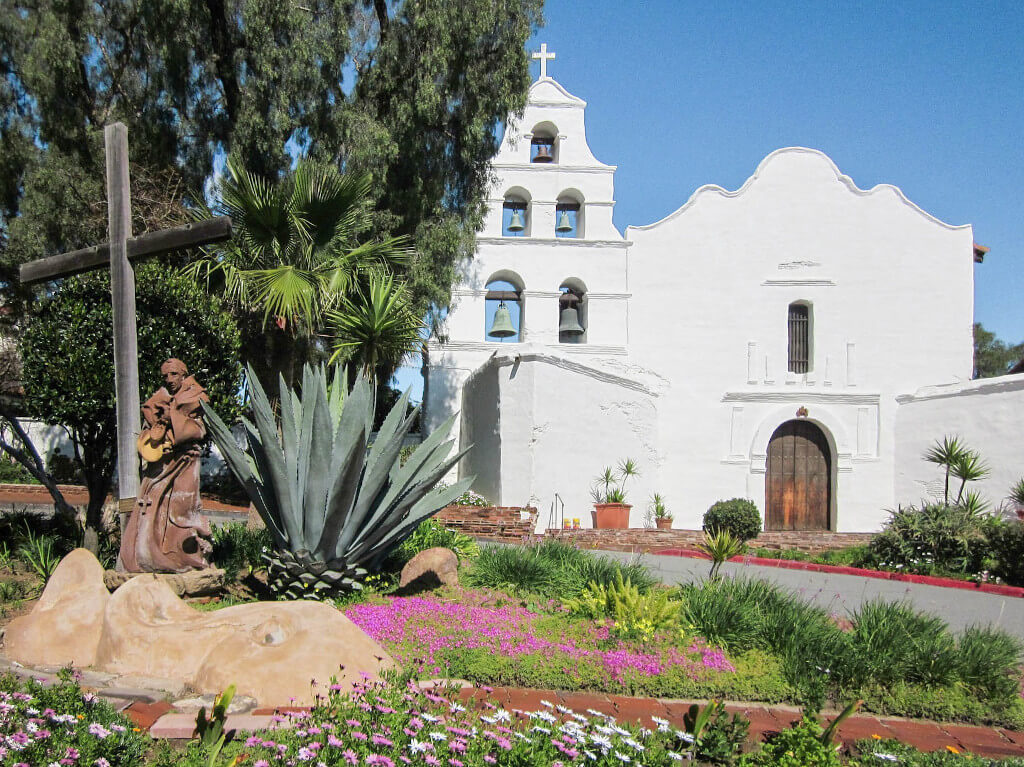 White exterior of the Mission Basilica San Diego de Alcala, with flowers and cross in front